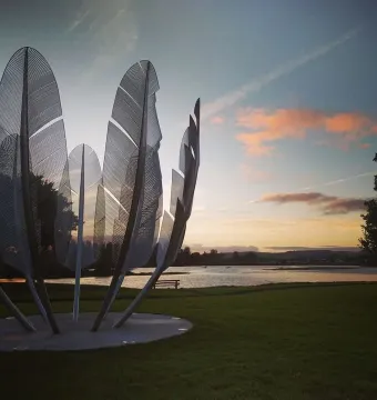 Kindred Spirits sculpture with curved metallic feather-like forms beside the water at sunset.