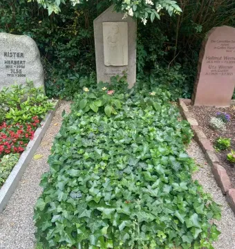Mourning Athena stone relief on a vertical headstone beside a small plaque and greenery.