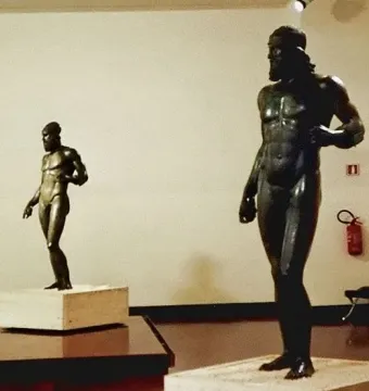 Two Riace bronzes on pedestals in a gallery, with one larger bearded nude male statue in the foreground and a smaller figure behind it.