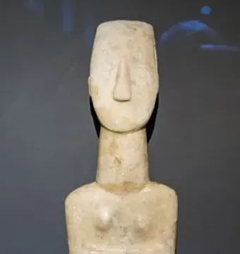 Kykladic idol with folded arms and a long, narrow marble body against a dark background.
