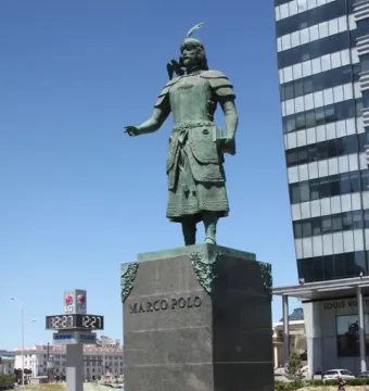 Marco Polo statue on a stone pedestal, with a bronze figure holding a book under a blue sky.
