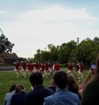 The Marine Corps War Memorial stands under a clear sky as a marching band in red colonial-style jackets performs on the lawn in front of it, with spectators nearby.