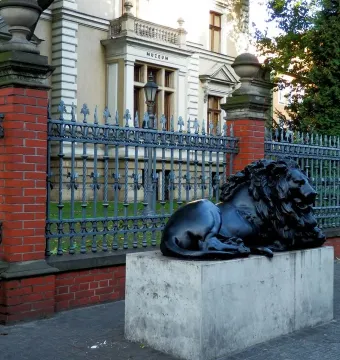 Guarding Lion sculpture on a light-colored pedestal beside an iron fence and brick wall, with a museum building in the background.