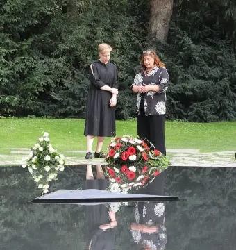 Two women stand beside the Memorial to the Murdered Sinti and Roma of Europe, next to a dark reflective water basin with floral wreaths.