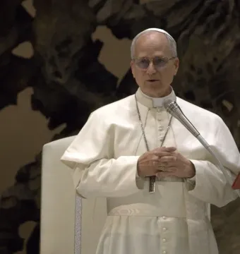 A man in white papal attire stands at a microphone with his hands clasped before a dark textured wall.