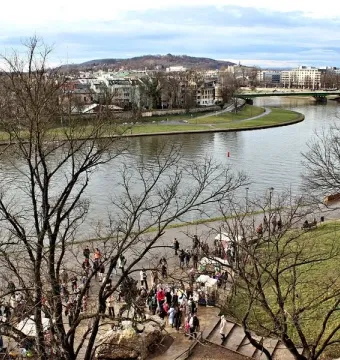 Wide view of the Vistula River with a cityscape, a riverside path full of people, and bare trees in the foreground.