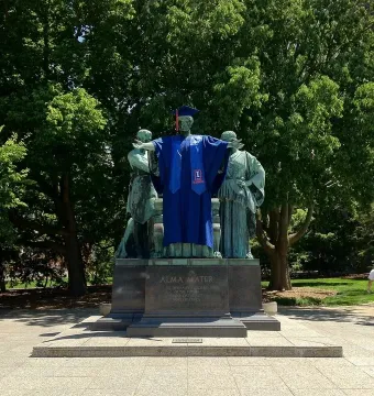The Alma Mater statue with a central bronze figure in a blue graduation gown, flanked by two figures on a stone pedestal.