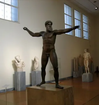 Artemision Bronze statue of a nude, bearded man with outstretched arms in a museum gallery.