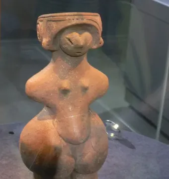Jōmon Venus clay figurine with a headdress, slit eyes, and rounded body on a dark display base.