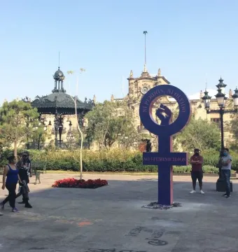 Antimonumenta, a purple pillar-like sculpture with a circular loop and raised fist, stands in a public square with pedestrians and a historic building in the background.