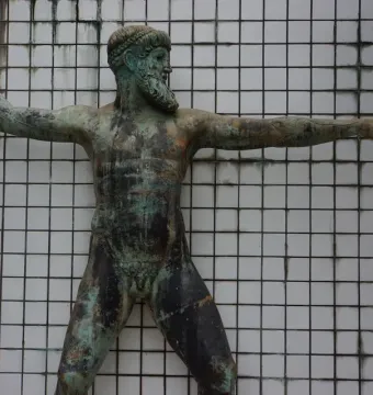 Artemision Bronze sculpture of a nude, bearded male figure with raised arm and extended arm, shown against a white tiled wall.