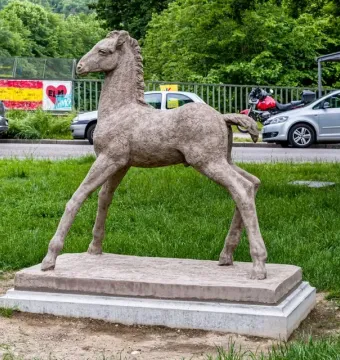 Holbeinpferd foal sculpture stands on a concrete base in grass, with a road, parked vehicles, graffiti-covered fence, and trees behind it.