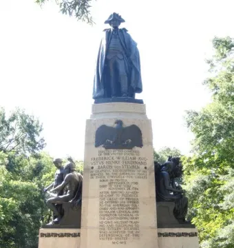 Von Steuben monument with a dark statue atop a stone pedestal, flanked by bronze figures and carved inscriptions.