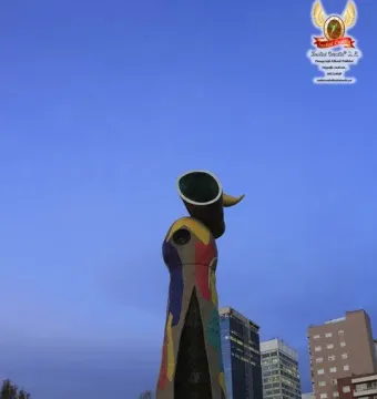 Dona i Ocell sculpture with colorful mosaic tiles and a tall vertical opening against a twilight city skyline.