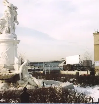 Lorelei Fountain in a snow-dusted park with an ornate stone basin and sculpture, with city buildings and a large screen behind it.