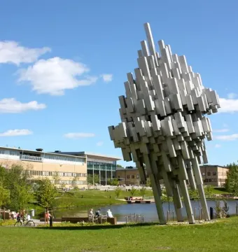 Norra skenet, a large abstract silvery sculpture on a lawn near a pond and modern campus buildings.