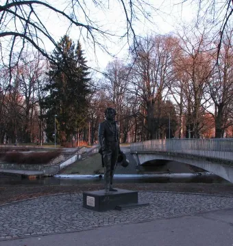 Pushkin statue on a dark pedestal in a park with bare trees and a bridge in the background