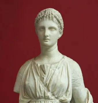 Artemis Diadoumena marble torso with draped folds and braided hair against a red background.