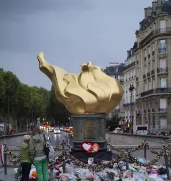 The Flame of Liberty sculpture stands above a memorial of flowers, photos, and locks, with two people nearby and Paris buildings in the background.