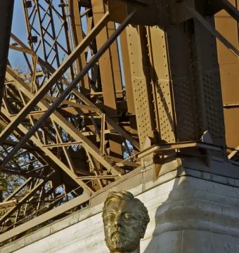 Gilded bust of Gustave Eiffel on a stone pedestal beneath the Eiffel Tower\'s metal framework.