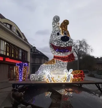 Reksio Monument lit with white and gold lights, with a red scarf and glowing gift boxes nearby.