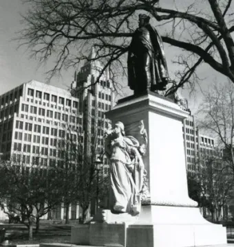 Commodore John Barry statue on a tall pedestal with a relief figure, shown in a park with bare trees and city buildings behind it.