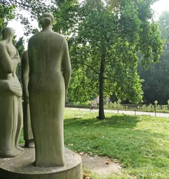 Three Standing Figures sculpture by Henry Moore on a circular base in a grassy park, seen from behind and the side.
