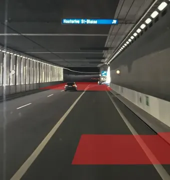 Interior of a brightly lit highway tunnel with a dark car ahead, white lane markings, an overhead Hauterive-St-Blaise sign, and red rectangular overlays on the road.