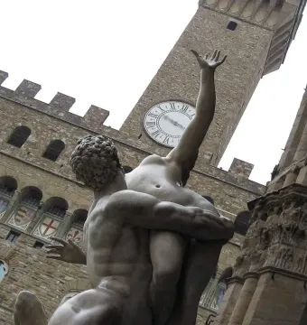Rape of the Sabine Women sculpture seen from below, with a muscular male figure lifting a nude woman in front of a clock tower.