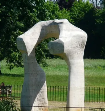 The Arch, a tall pale abstract stone sculpture in a grassy park with a low fence and trees behind it.