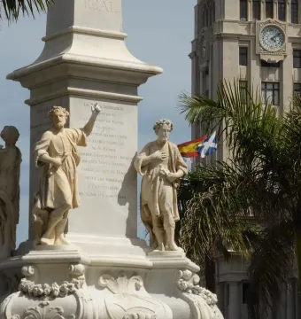 Triunfo de la Candelaria monument with carved figures on a pedestal, with a clock tower, palm trees, and flags in the background.