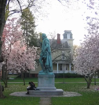 William the Silent statue on a stone base in a grassy area with pink magnolia trees and a yellow building behind it.