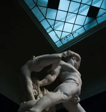 Hercules and Lychas marble sculpture lit against a dark background with a blue skylight above.