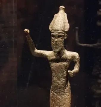 Dark metal statuette of Melqart di Sciacca with a pointed headdress and raised arm, displayed on a light base inside a case.