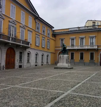 Monument to Mosè Bianchi, a dark seated statue on a tall pedestal in a cobblestone piazza.