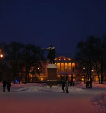 Pushkin statue with an outstretched arm in a snow-covered park at night, with an illuminated building behind it.