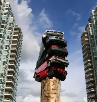 Trans Am Rapture sculpture with stacked cars mounted on a tall stripped tree trunk in Vancouver, framed by high-rise buildings.
