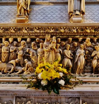 Intricate relief sculpture on the Arca di San Domenico, showing multiple carved figures around a Madonna and Child scene, with flowers in the foreground.