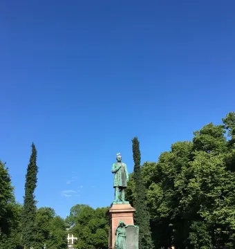 Johan Ludvig Runeberg statue on a stone pedestal with trees and a bright blue sky behind it, topped by a white bird.
