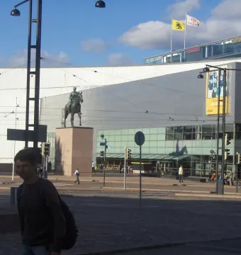 Marshal Mannerheim equestrian statue in front of a modern glass building in Helsinki, with a pedestrian in the foreground.