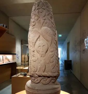 Pfalzfeld Obelisk, a carved sandstone monument with an anthropomorphic face and swirling patterns, displayed on an orange pedestal in a museum corridor.