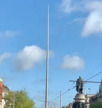 The Spire towers over a busy Dublin street with buses, cars, pedestrians, and surrounding buildings under a blue sky.
