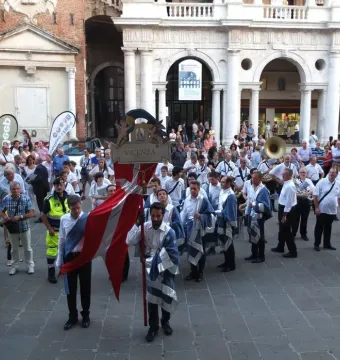 A crowd gathers in a paved square beside an ornate colonnaded building, with a red-and-white Vicenza banner held at the center and musicians in blue-and-white garments nearby.