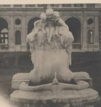 Stone monument with three seated figures, known as Children and Frog, standing on a lawn with an ornate building behind it.