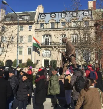 Mahatma Gandhi statue with a crowd gathered nearby in front of a building, with the Indian flag flying in the background.