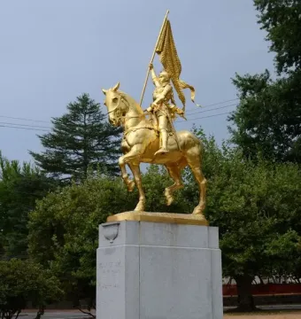 Gilded equestrian statue of Joan of Arc in armor holding a banner on a stone pedestal.
