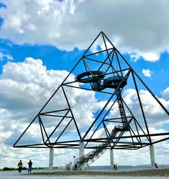 The Tetrahedron steel sculpture stands on tall white pillars with staircases and viewing platforms, with people walking nearby under a blue sky.