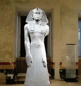 Statue of Amenemhat III standing upright in a museum gallery, wearing a striped nemes headdress and a flared kilt.