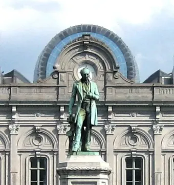 John Cockerill Monument with a standing bronze figure on an ornate pedestal and two seated figures below, in front of a grey stone building.