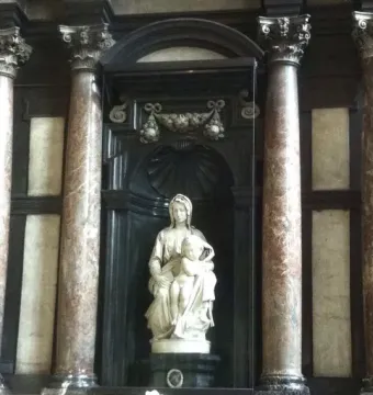 Madonna of Bruges marble sculpture seated with the Christ Child in a dark carved niche behind glass.